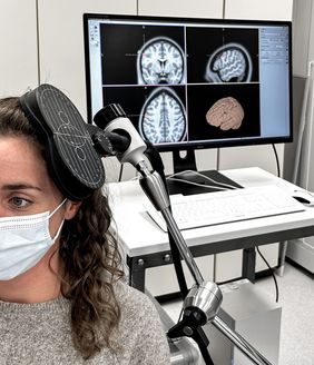 Frau hat eine Spule am Kopf, von der aus gezielt magnetische Impulse durch die Schädeldecke (transkraniell) an bestimmte Hirnareale abgegeben werden, um deren Aktivität zu beeinflussen. Im Hintergrund ein Monitor mit Bildern vom Gehirn. 