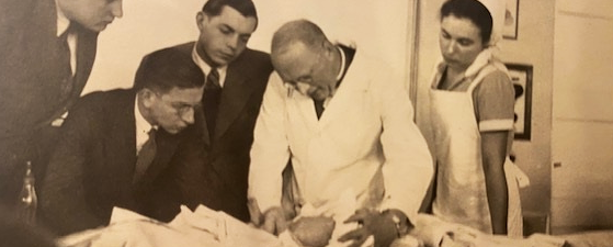 Das Bild zeigt Hans Rietschel, der im weißen Kittel einen Säugling untersucht, daneben eine SChwester in Schwestertracht, links schauen drei Studierende in dunklen Anzügen zu.