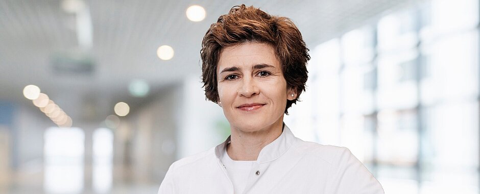Prof. Dr. Anke Katharina Bergmann leitet das im Oktober 2025 gegründete Institut für Klinische Genetik und Genommedizin Anke Bergmann steht in weißem Kittel in einem Flur, der Hintergrund ist sehr unscharf. Das Bild ist im Querformat, Frau Bergmann ist bis zum Bauchnabel zu sehen.