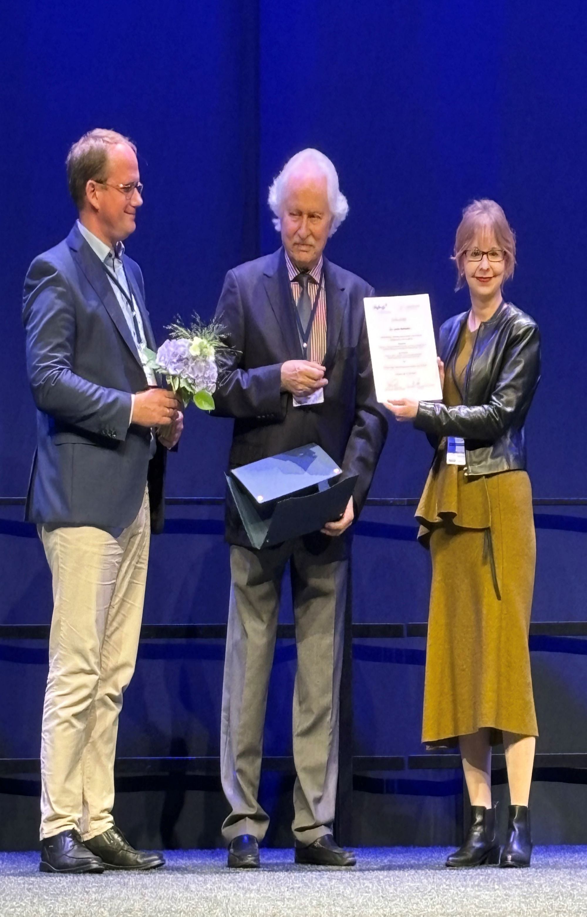 Laudator Tobias Renner hält auf der Bühne einen Blumenstrauß in den Händen, daneben Hans-Christoph Steinhausen, der Julia Geissler die Urkunde überreicht. 