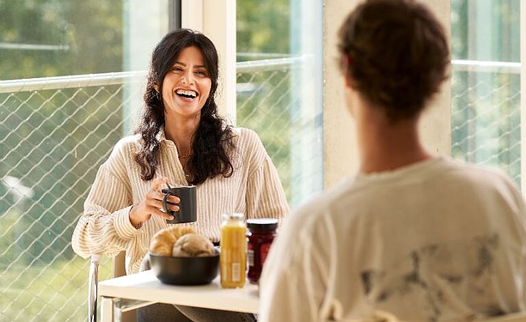 FLEX4UKW Café © Mirko Nyssen Person sitzt an einem Frühstückstisch, lächelt in die Kamera und hält einen Kaffee in der Hand