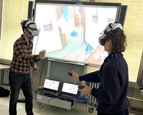 Zwei Personen mit VR-Brillen auf dem Kopf und Sticks in der Hand, im Hintergrund ist das virtuelle Szenario auf dem Monitor angezeigt. 