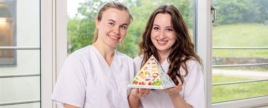 Zur Ausbildung Diätassistenz Symbolbild Diätassistenz: Zwei Auszubildende halten das Modell einer Ernährungspyramide hoch