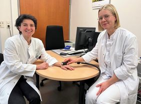 Nurcan Üceyler und Betty Feulner sitzen in weißen Kitteln am Besprechungstisch - Betty Feulner hält ein Messgerät auf die Hand von Nurcan Üceyler. 