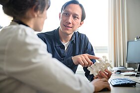 Johannes Tran-Gia hält in der Hand ein 3D-gedrucktes Modell der Lendenwirbelsäule, zeigt mit der anderen Hand darauf und schaut seine Kollegin links im Bild an. 