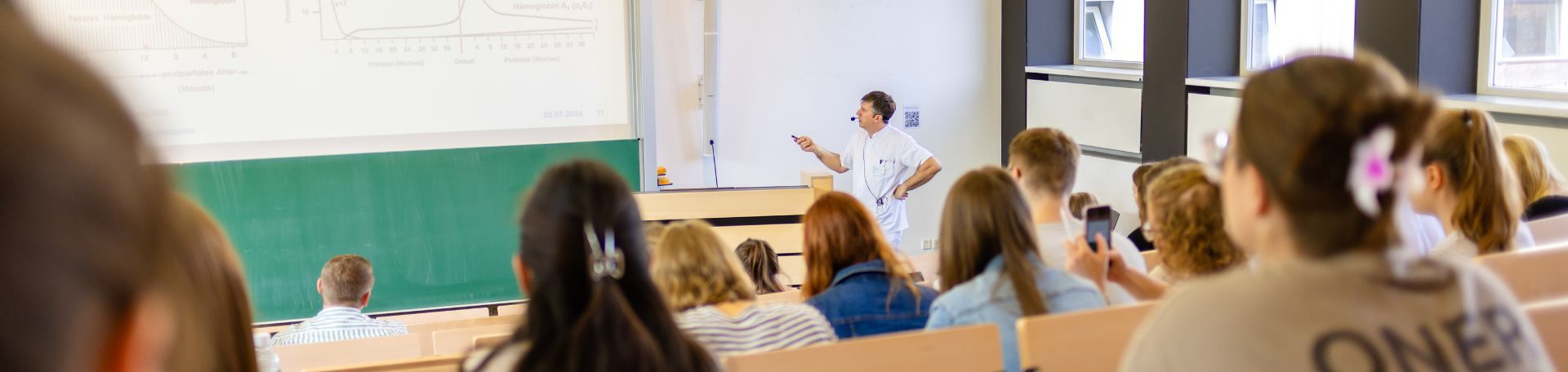 Ein Dozent erläutert vor Studierenden in einem Hörsaal eine Graphik