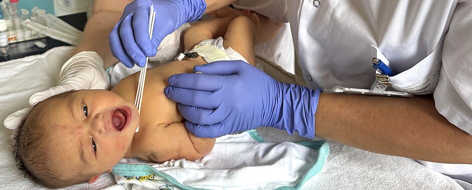 MIAI - Hautabstrich Säugling liegt auf Wickelkommode, Studienärztin nimmt mit Wattepad einen Abstrich von der Haut, im Hintergrund steht die Studynurse mit Mundschutz