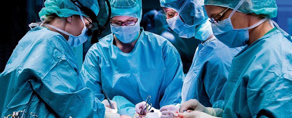 4 Fachkräfte in OP-Kluft mit Kittel, Haube und Mundschutz stehen am Operationstisch und behandeln einen Patienten