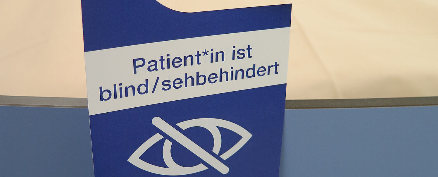 Auf dem Bild sieht man ein frisch bezogenes Krankenbett an dem am Ende des Fußes ein blauer Anhänger am Gestänge hängt. Auf dem steht Patient*in ist blind / sehbehindert. Darunter sind zwei Piktogramme. Ein durchgestrichenes Auge und ein Strichmännchen mit Stock, der vor einer Stufe steht.