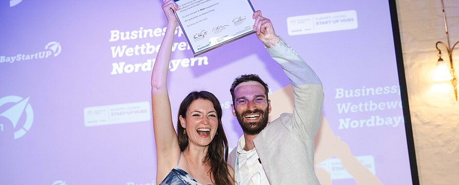 ENDOLEASE Anna Fleischer und Johannes Braig Anna Fleischer im blau-weißen Sommerkleid und Johannes Braig im beigefarbenen Jacket halten auf der Bühne die eingerahmte Urkunde hoch und strahlen in die Kamera.