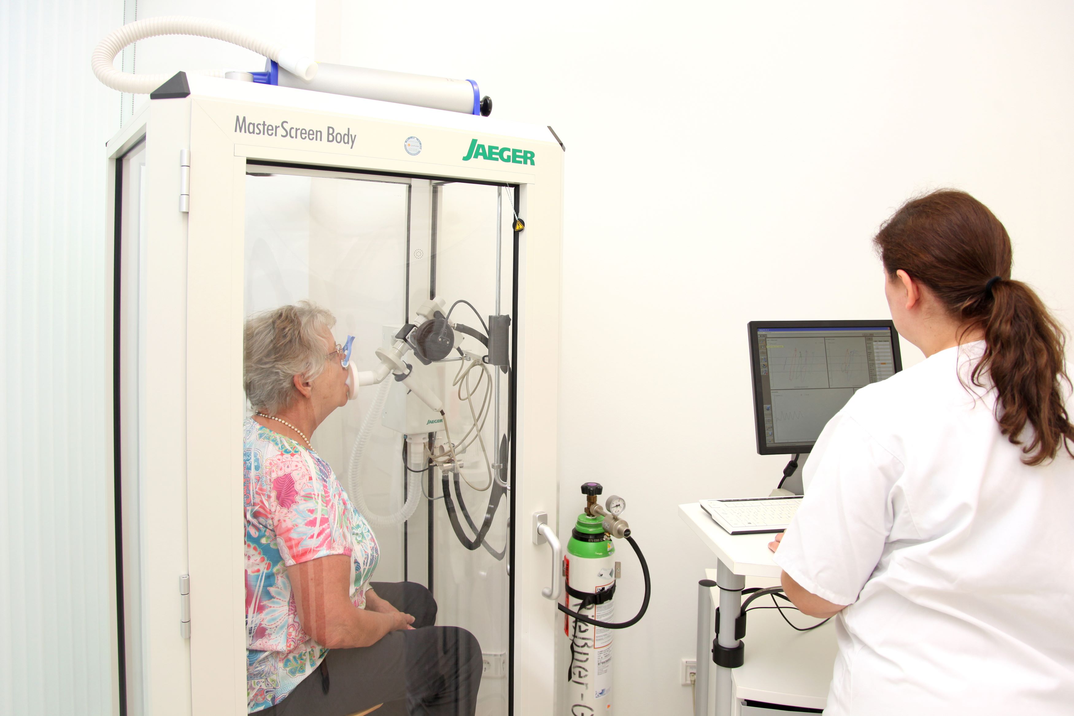 Eine Patientin sitzt in einer Kabine mit Maske vor dem Mund, eine Nurse leitet sie an, kräftig auszuatmen.n. 