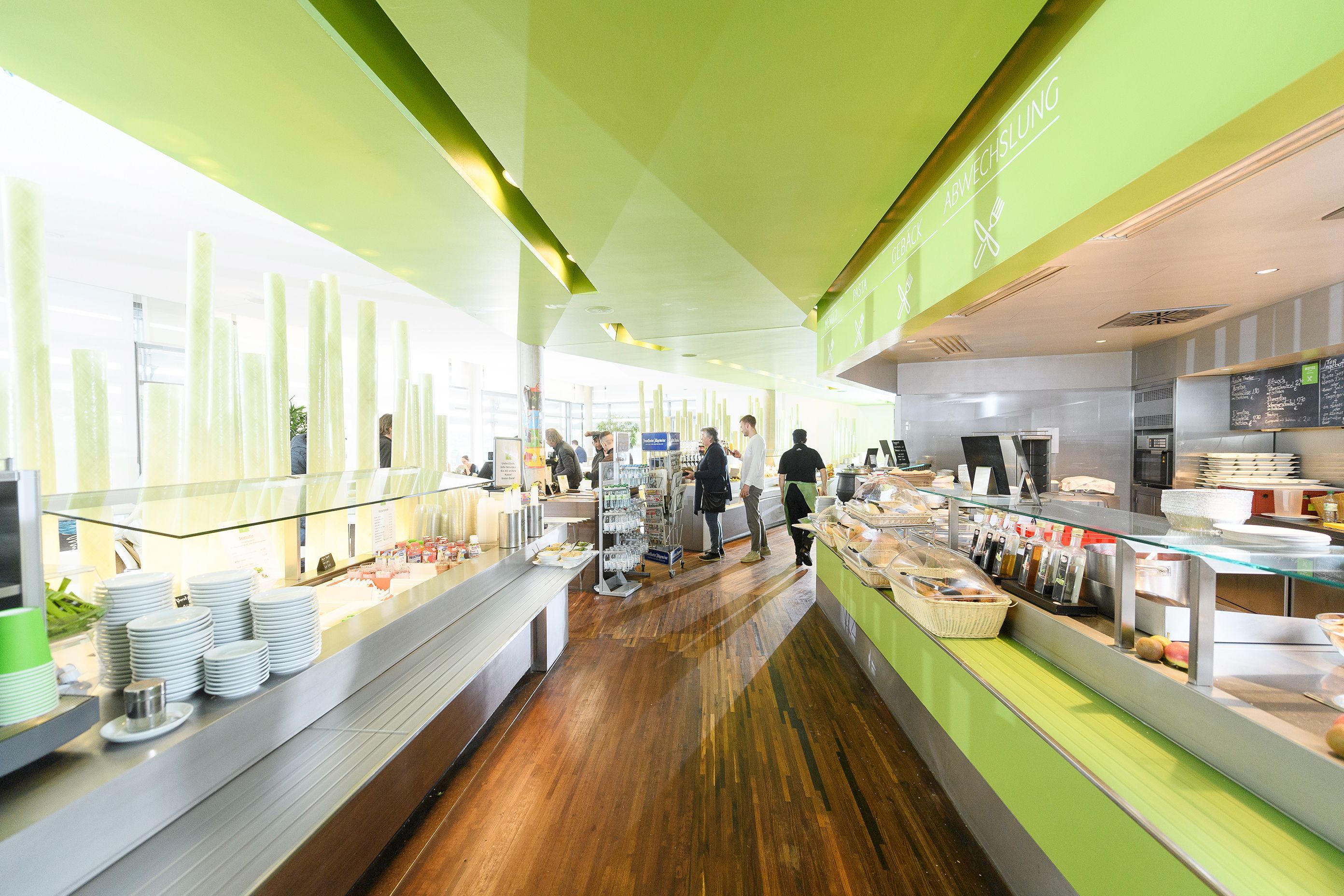 Blick in das UKW-Restaurant, Selbstbedienungstheken rechts und links, in frischer grüner Thekenverkleidung, grüner Decke und dunklem Holzstrukturboden.