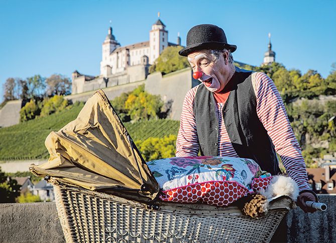 Clown Jojo schaut in einen altertümlichen Requisiten-Kinderwagen hinein. Er steht auf einer Steinbrücke, im Hintergrund die Würzburger Festung Marienburg.