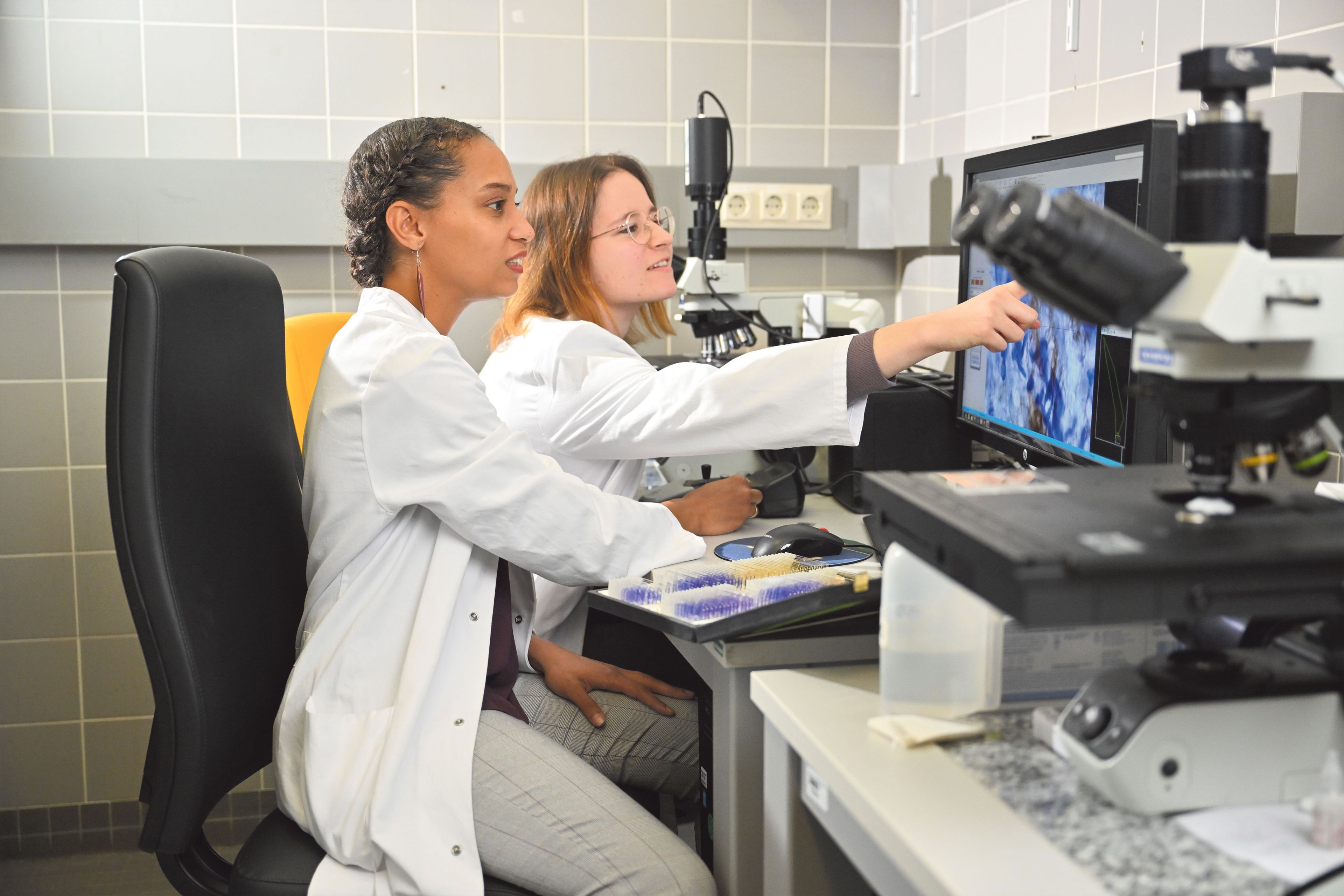 Frau Prof. McFleder sitzt neben einer Kollegin im Labor an einem Elektronenmikroskop, dessen Objektträger-Aufnahme auf einem Computerbildschirm angezeigt wird.