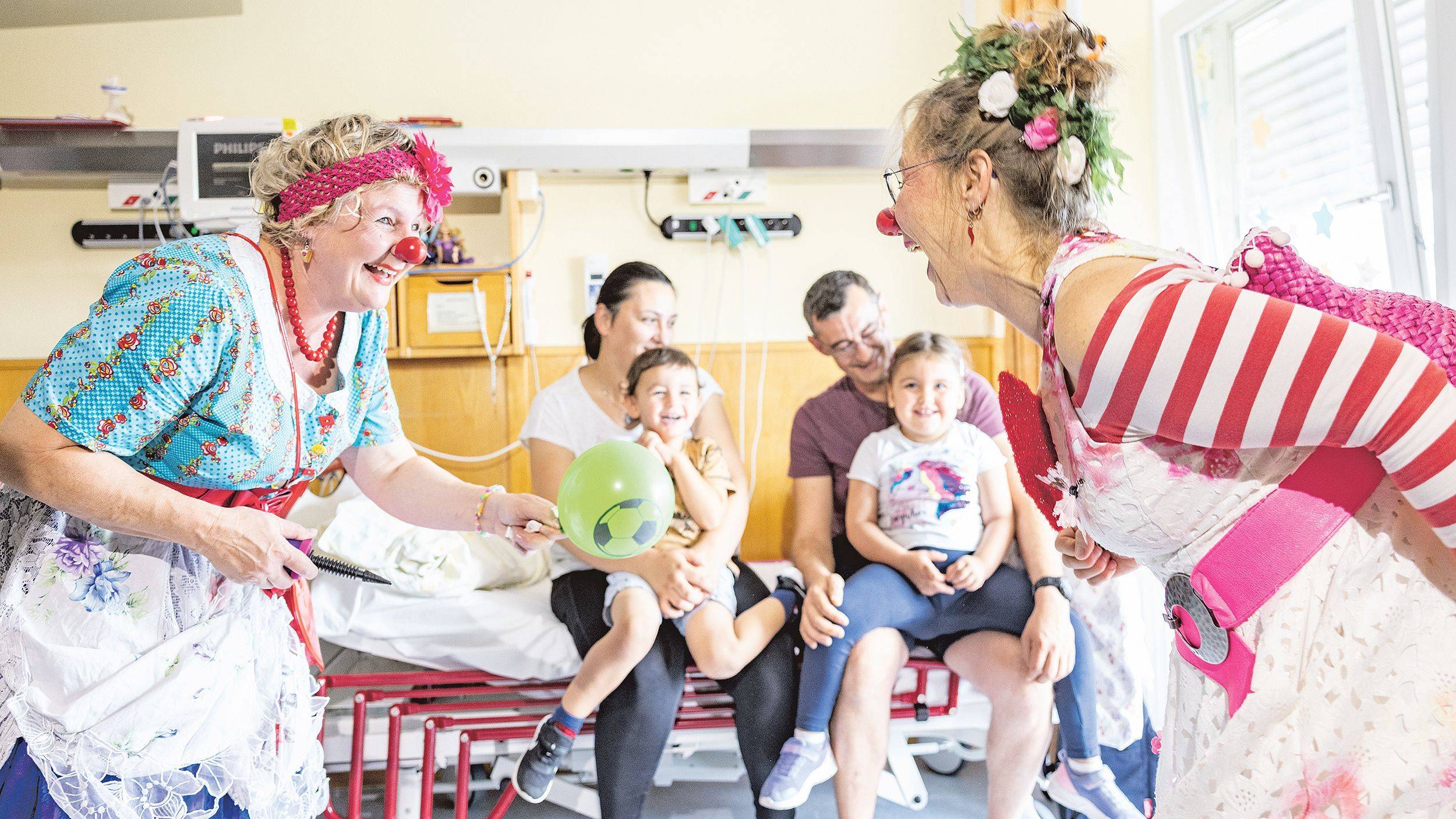Schlawine und Flora bespaßen in einem Patientenzimmer eine Familie mit zwei lachenden, kleinen Kindern.