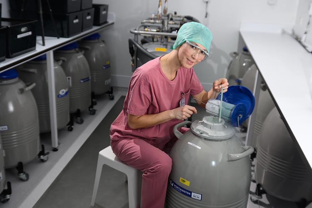 : Frau in Laborkleidung sitzt vor einem geöffneten Kryotank, in dem Eizellen und Spermien eingefroren sind.