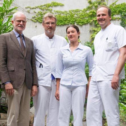 Gruppenbild: Vier Menschen stehen vor einer Efeu-bewachsenen Wand, drei Männer und eine Frau. Einer von ihnen trägt einen Anzug, drei tragen Arztkleidung.