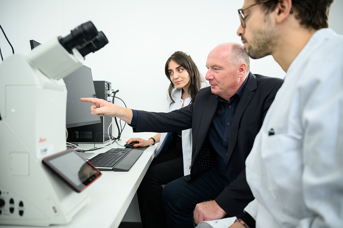 Prof. Nieswandt sitzt vor einem Computerbildschirm und zeigt darauf mit dem Zeigefinger. Neben ihm sitzen eine Frau und ein Mann im Laborkittel und schauen auf den Bildschirm. Links im Bild ist ein Mikroskop zu sehen.