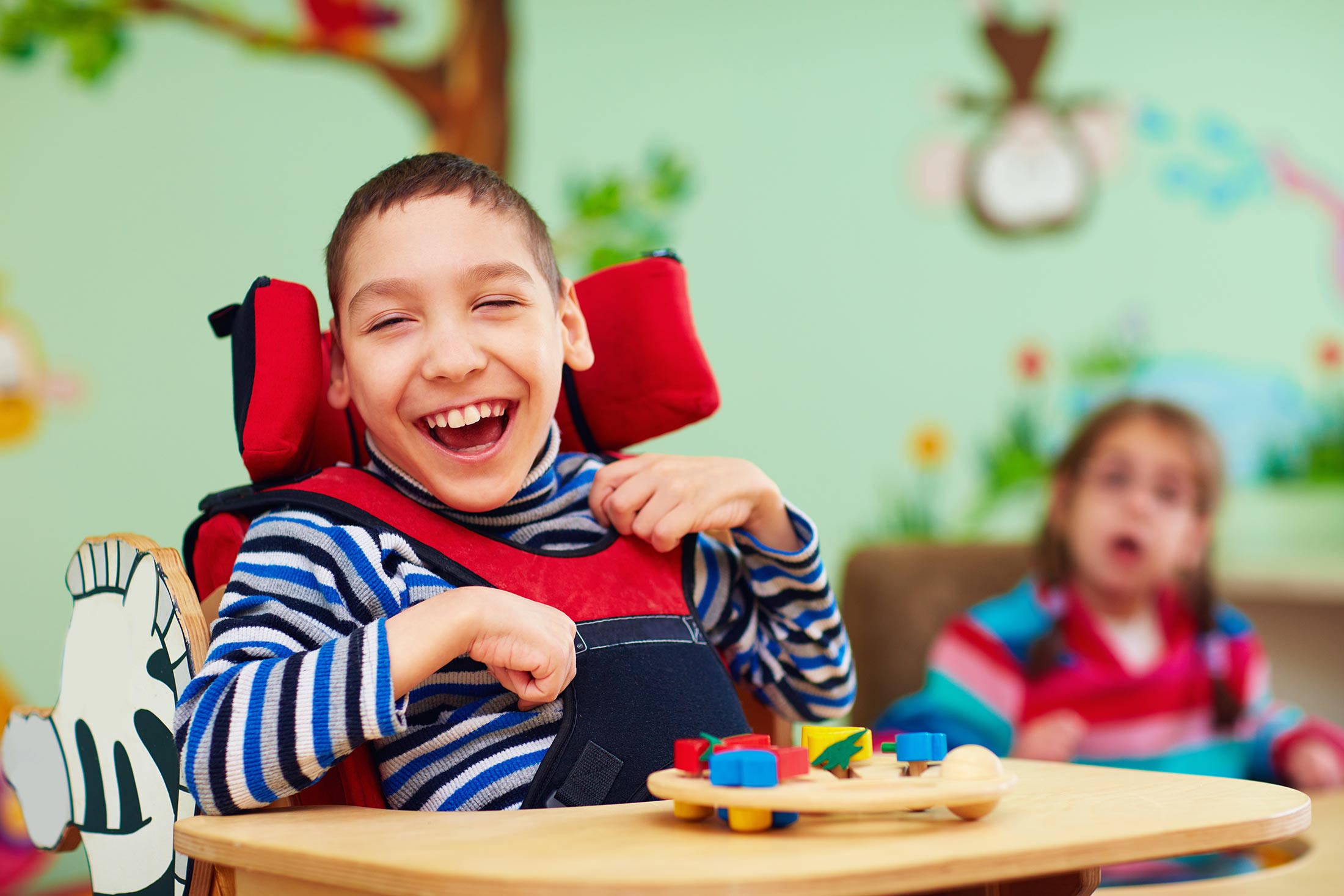 Das Bild zeigt einen fröhlichen Jungen mit Behinderung, der in einem roten Rollstuhl sitzt und mit einem bunten Holzspielzeug spielt. Der Junge lächelt breit und wirkt glücklich und zufrieden. Im Hintergrund ist unscharf ein weiteres Kind zu sehen.