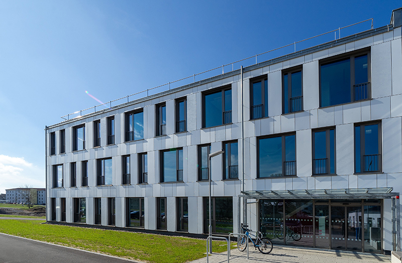 Das Bild zeigt die Fassade eines grauen, modernen Gebäudes mit vielen Fenstern, im Hintergrund strahlend blauer Himmel.