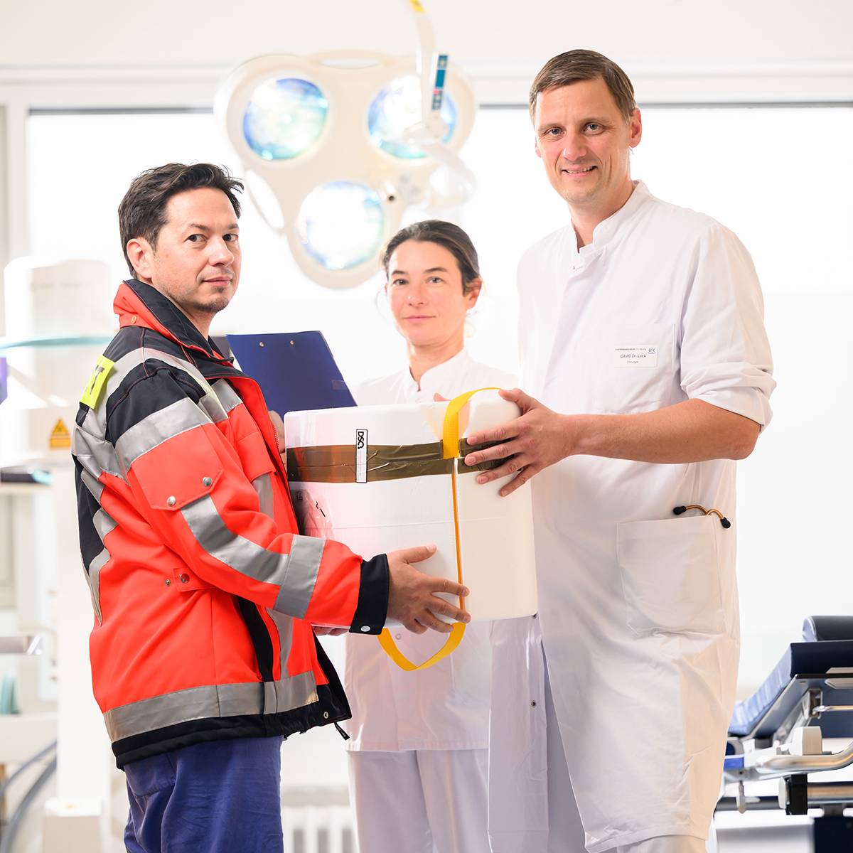 Das Bild zeigt drei Personen in medizinischem Umfeld. Eine Person in roter Rettungsdienstjacke übergibt ein weißes Paket an einen Mann im Arztkittel. Im Hintergrund sieht man eine Frau in weißer Kleidung und eine Operationsleuchte.