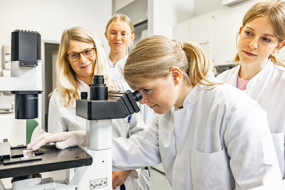 Eine junge Frau schaut durch ein Mikroskop. Hinter ihr stehen zwei weitere junge Frauen, die sie interessiert beobachten. Neben ihr steht Prof. Stefanie Hahner und lächelt. Alle tragen Arztkittel.