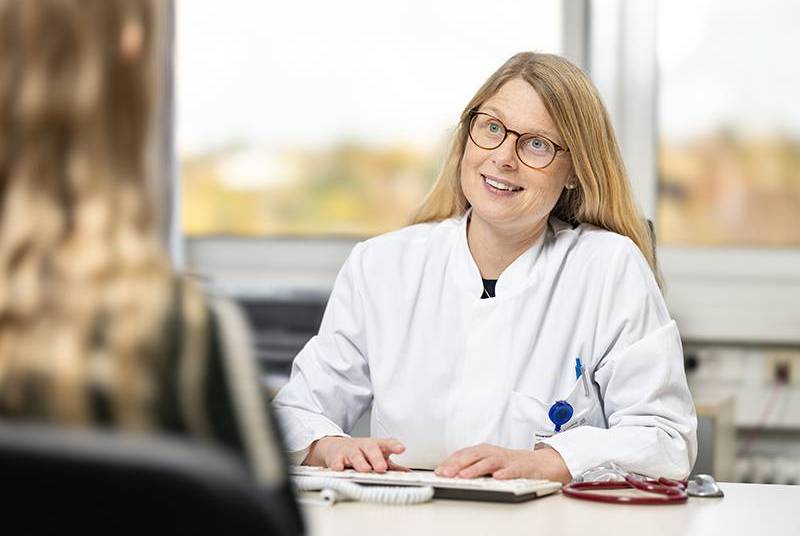 Prof. Stefanie Hahner sitzt an einem Schreibtisch, vor ihr eine Tastatur und ein Stethoskop. Sie spricht mit einer Frau, die vor ihr sitzt und die unscharf von hinten zu sehen ist.